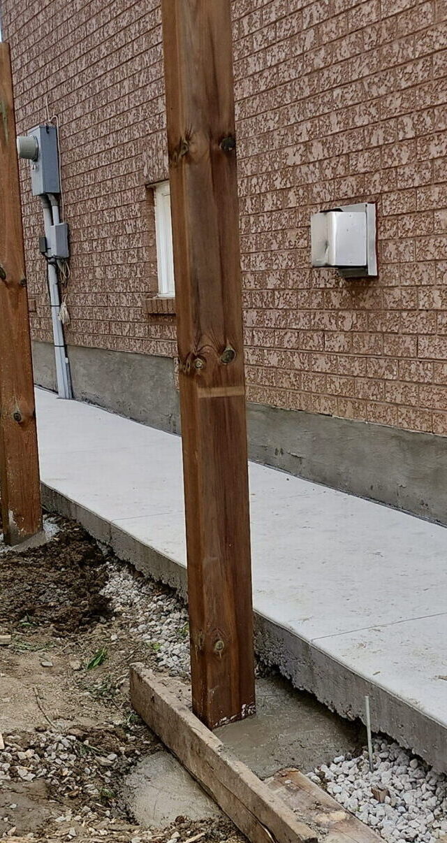 Wood fence post set in concrete footing beside house with gravel drainage and walkway